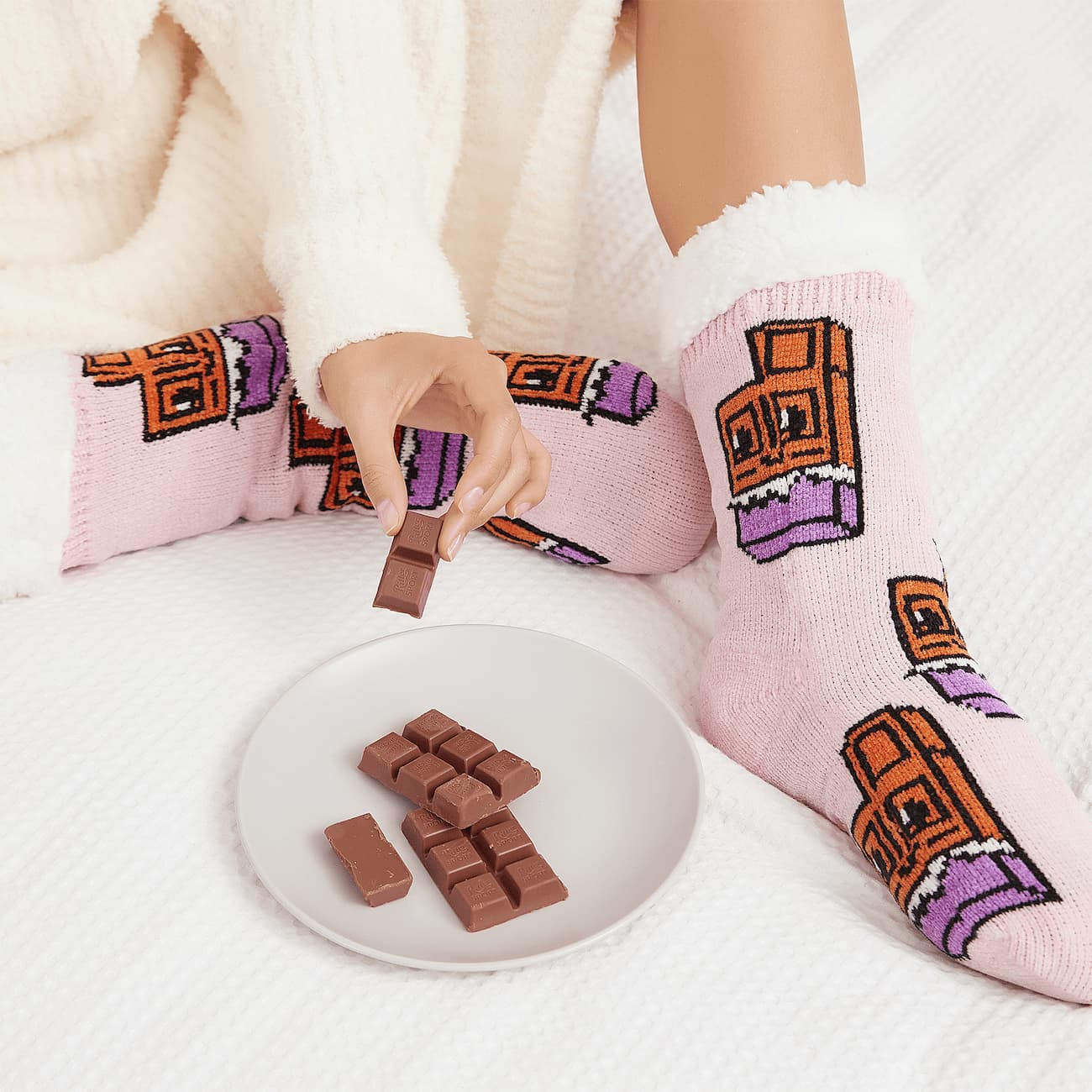 The Foozie™ Chocolate slipper socks, worn on a white bed beside a plate of chocolate